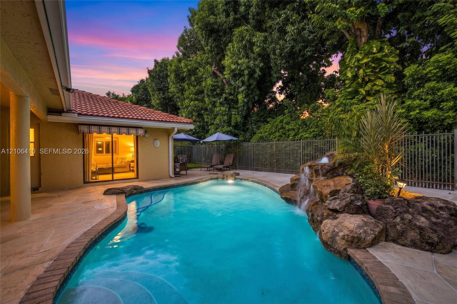 1725 Lakeshore Circle, Weston, FL 33326 In-ground swimming pool featuring a naturalistic rock waterfall, stone-tile decking, and surrounding metal fencing against a backdrop of mature trees