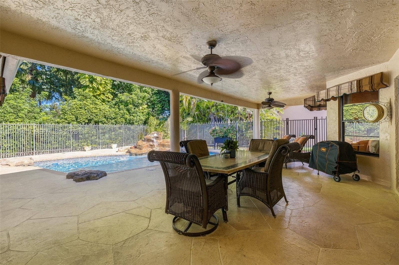 1725 Lakeshore Circle, Weston, FL 33326 Covered outdoor patio featuring stone-patterned concrete flooring, two ceiling fans, a swimming pool with a rock water feature, and a metal perimeter fence