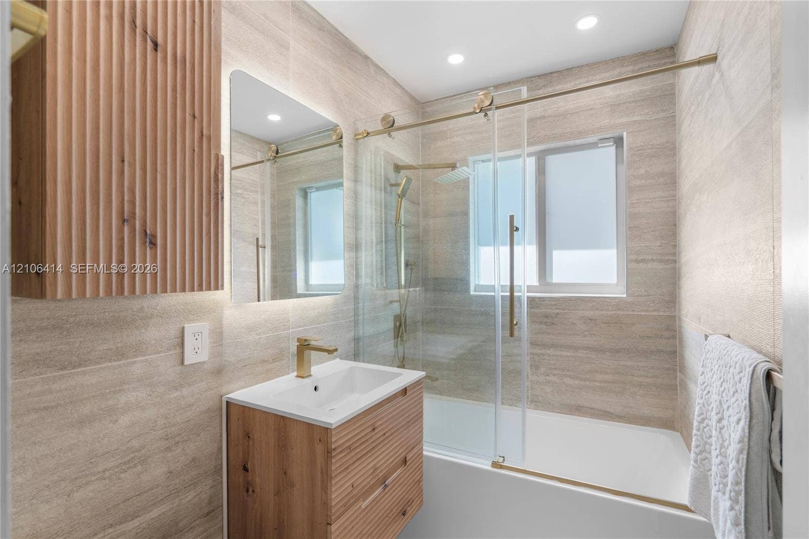 9133 Garland Avenue, Surfside, FL 33154 Bathroom featuring floor-to-ceiling stone-look tiling, a slatted wood-finish vanity with an integrated basin, gold-tone hardware, and a glass-enclosed bathtub