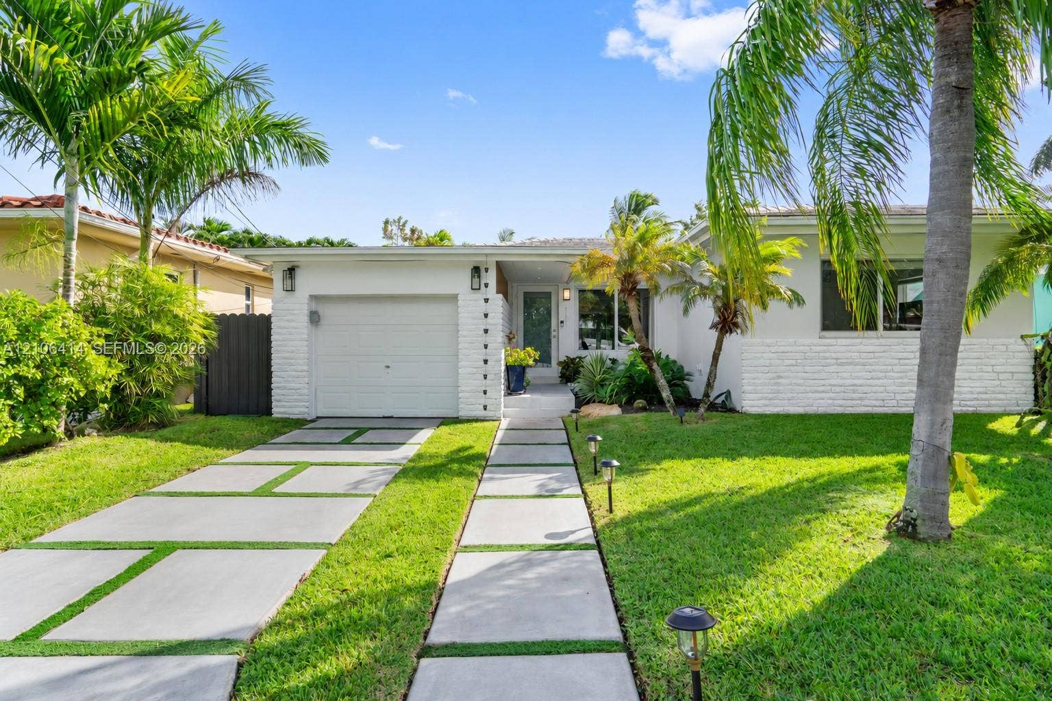 9133 Garland Avenue, Surfside, FL 33154 Single-story residence featuring white painted brick masonry, a concrete paver driveway, a manicured lawn, and mature palm trees