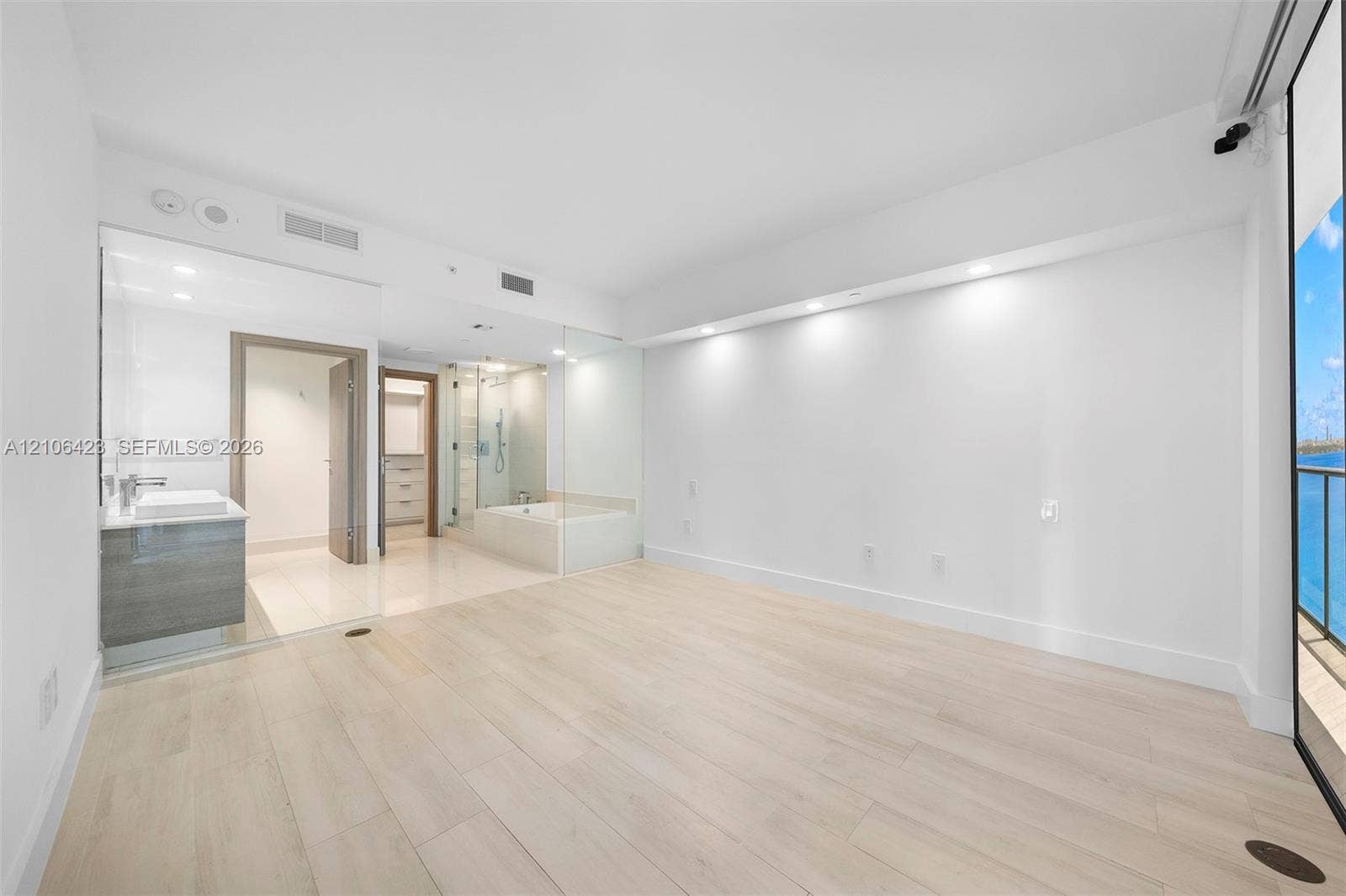 Open-concept space featuring light-toned wood-finish flooring, recessed architectural soffit lighting, glass-enclosed shower, soaking tub, and floor-to-ceiling glass panel balcony access