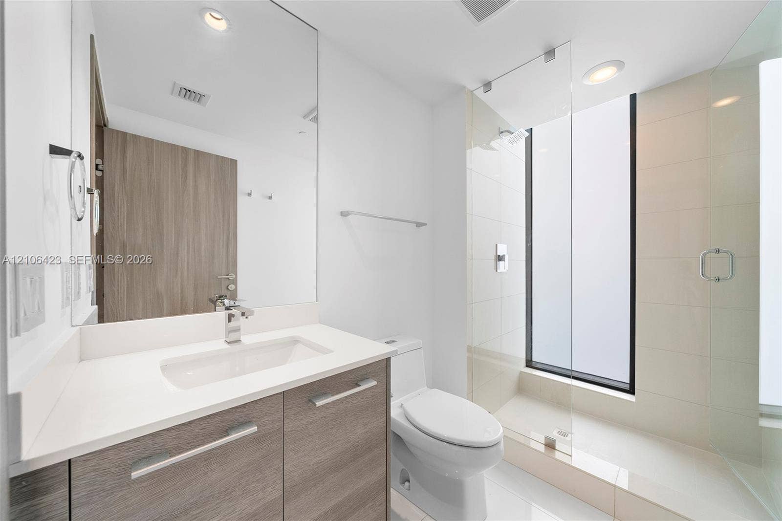 Bathroom featuring a floating wood-finish vanity with a white countertop, large frameless mirror, glass-enclosed walk-in shower with floor-to-ceiling tile, and recessed lighting
