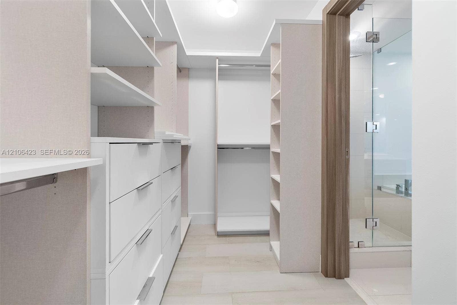 Walk-in closet featuring custom white cabinetry, open shelving, integrated drawer units, and tile flooring, including a glass-enclosed shower with wood-finish trim