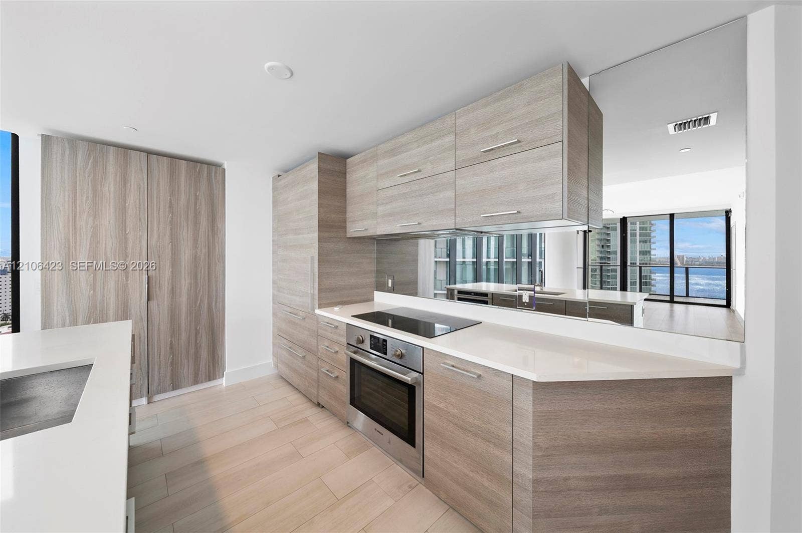 Modern kitchen featuring flat-panel wood-finish cabinetry, white solid-surface countertops, a mirrored backsplash, stainless steel built-in oven with electric cooktop, and light-toned wood-finish flooring