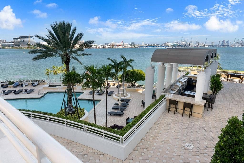 808 Brickell Key Drive, #2102, Miami, Miami, FL 33131 Waterfront pool terrace featuring a paved sun deck, built-in outdoor bar with a covered structure, mature palm trees, and perimeter landscape hedging