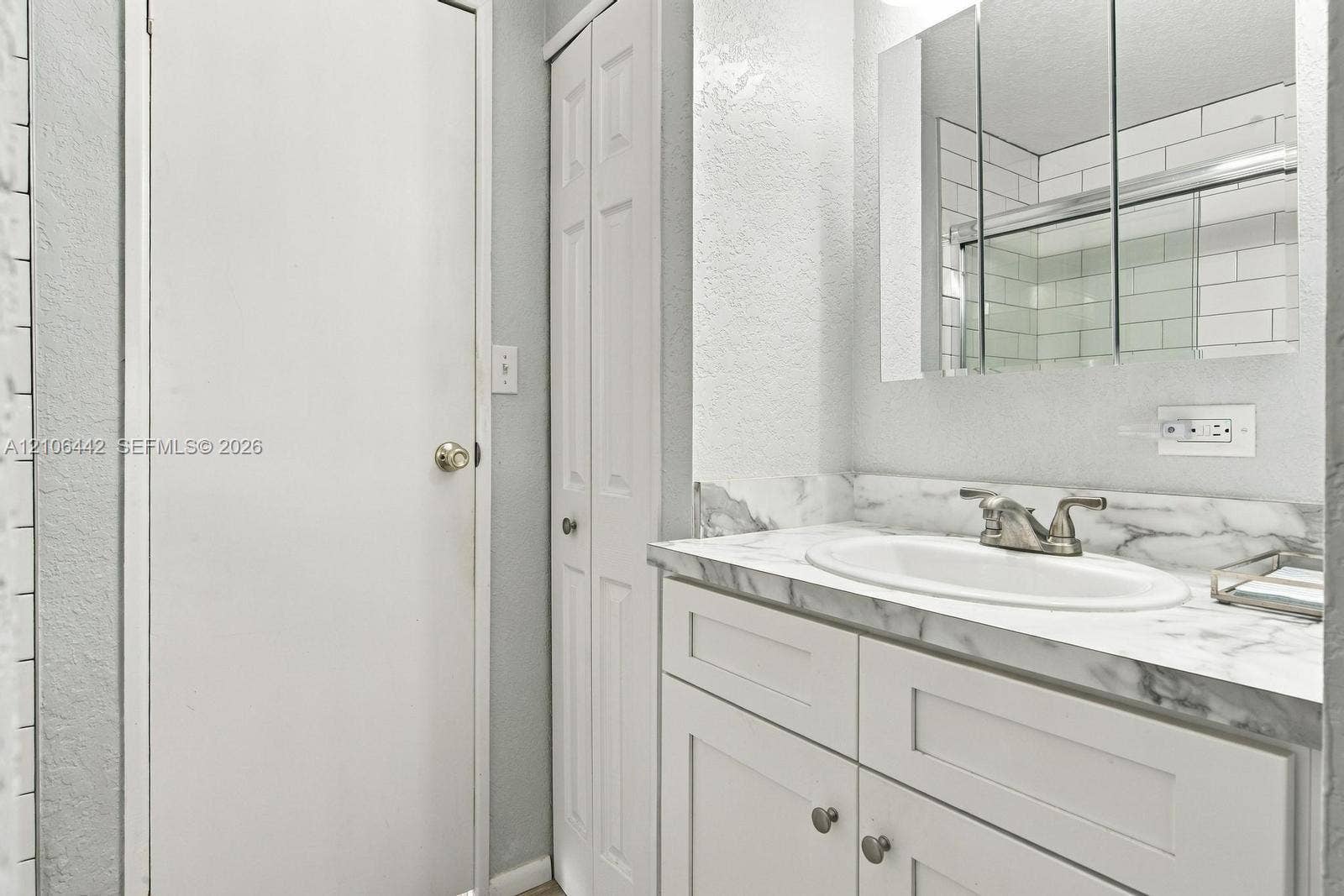 White vanity with stone-finish countertop, oval undermount sink, three-panel medicine cabinet, and tall storage closet with louvered doors