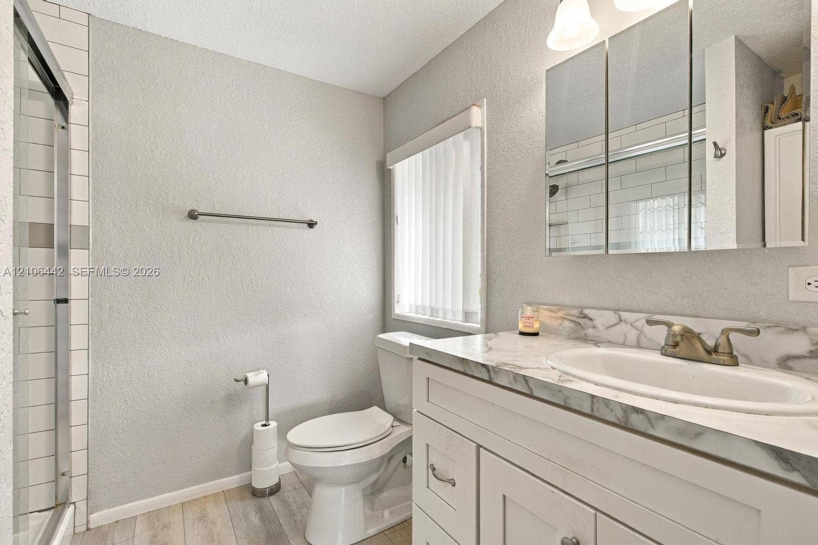 Bathroom featuring a white vanity with a patterned stone-finish countertop, integrated sink, three-panel medicine cabinet, tiled walk-in shower, and wood-finish flooring