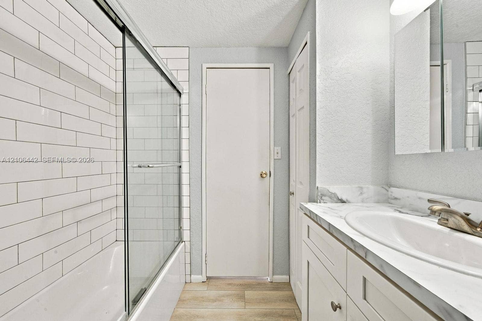 Bathroom featuring a tub-shower combination with white subway tile surround, sliding glass door, stone-finish vanity countertop with integrated basin, and wood-finish flooring