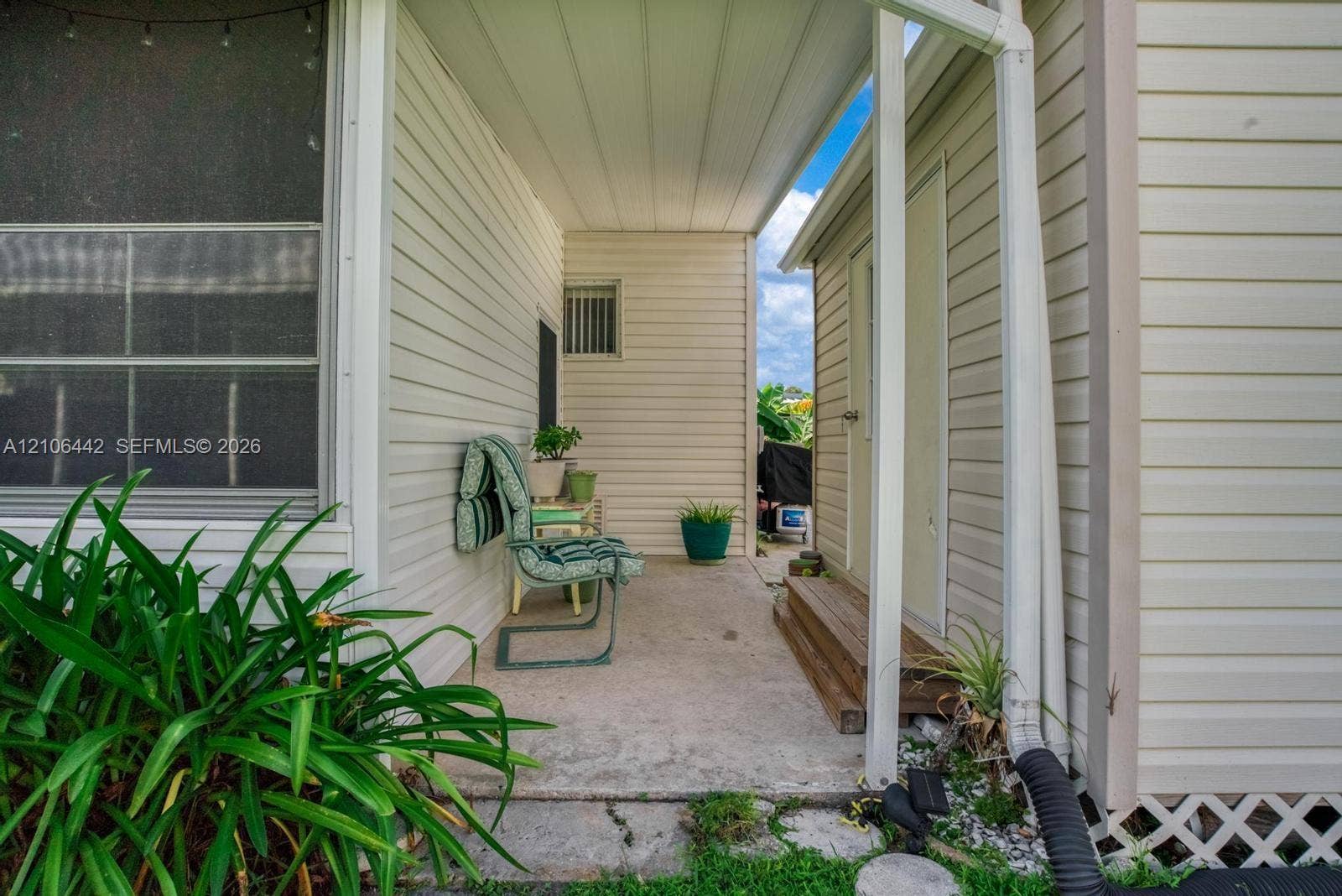 Covered exterior patio featuring light-colored horizontal siding, concrete flooring, a wooden two-step staircase, and white structural support columns