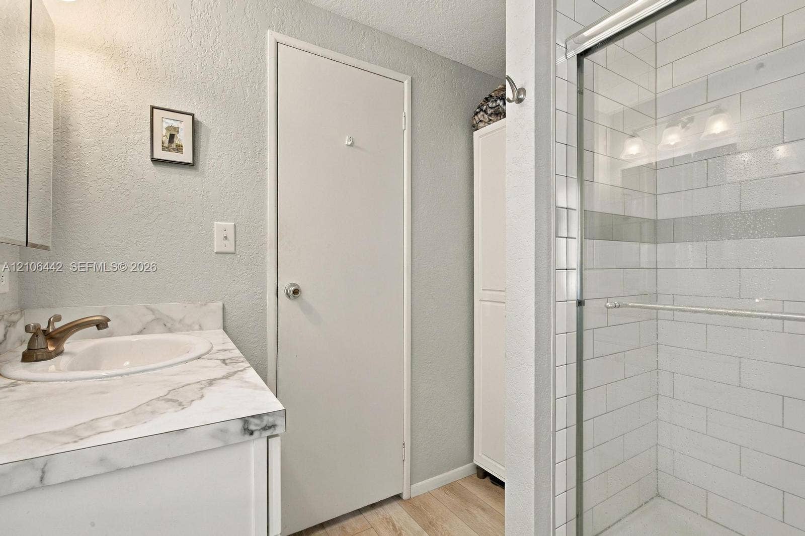 Renovated bathroom featuring a marble-topped vanity with integrated sink, polished brass faucet, subway tile shower enclosure with glass partition, wood-finish flooring, and soft ambient lighting from recessed fixtures