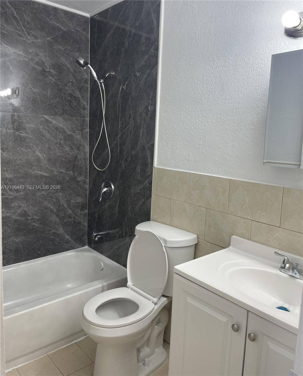 Bathroom featuring a shower-tub combination with dark patterned wall tile, a white vanity with an integrated basin, a polished chrome faucet, and neutral wall-mounted ceramic tile backsplashes