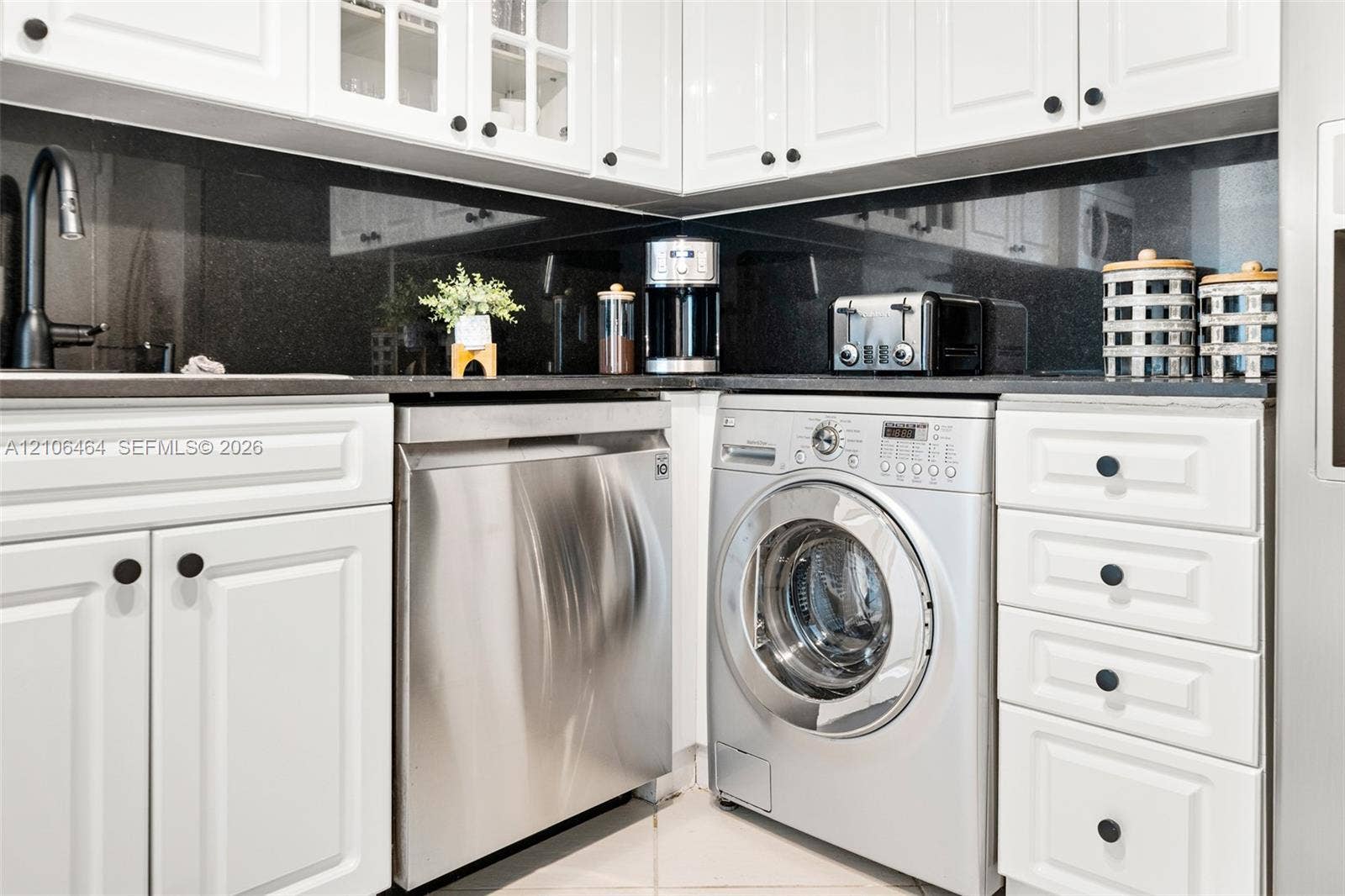 6770 Indian Creek Drive, #8E, Miami Beach, FL 33141 White cabinetry with raised-panel doors, solid-surface dark countertops, full-height dark backsplash, stainless steel dishwasher, and integrated front-loading washer