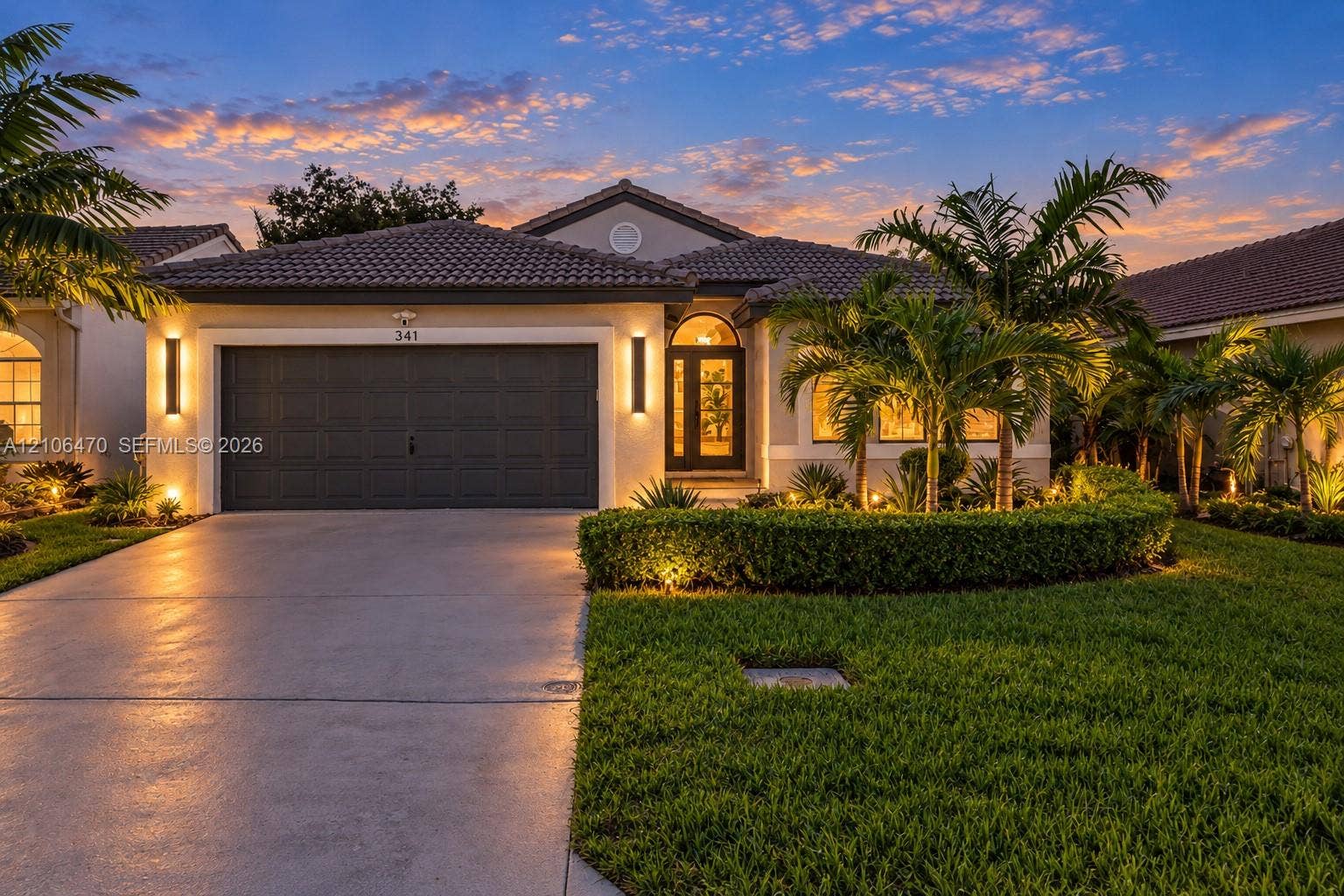 Single-story residence featuring a tile roof, concrete driveway, attached multi-car garage, manicured landscaping, and exterior wall-mounted lighting