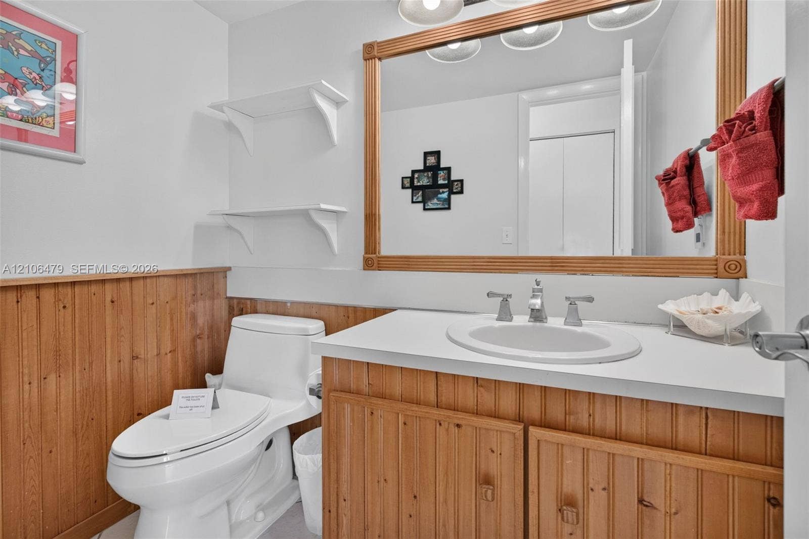 2800 SW 87th Avenue, #1113, Davie, FL 33328 Vanity featuring a solid surface countertop, integrated oval basin, and wood-finish cabinetry with wainscoting, accented by a large wood-framed mirror and wall-mounted shelving