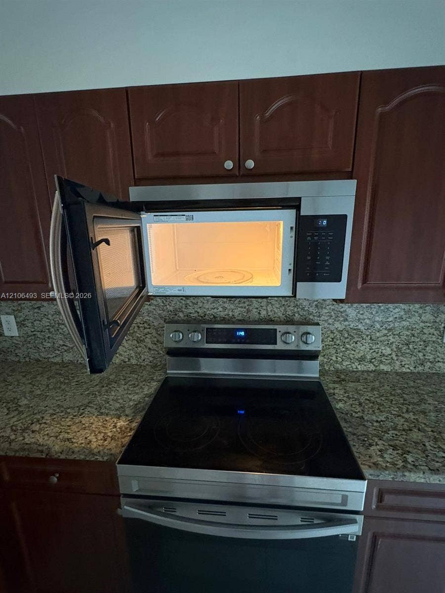 117 SE 3rd Avenue, #7, Hallandale Beach, FL 33009 Stainless steel over-the-range microwave and electric range, dark wood-finish cabinetry, and stone-surface countertops with matching backsplash