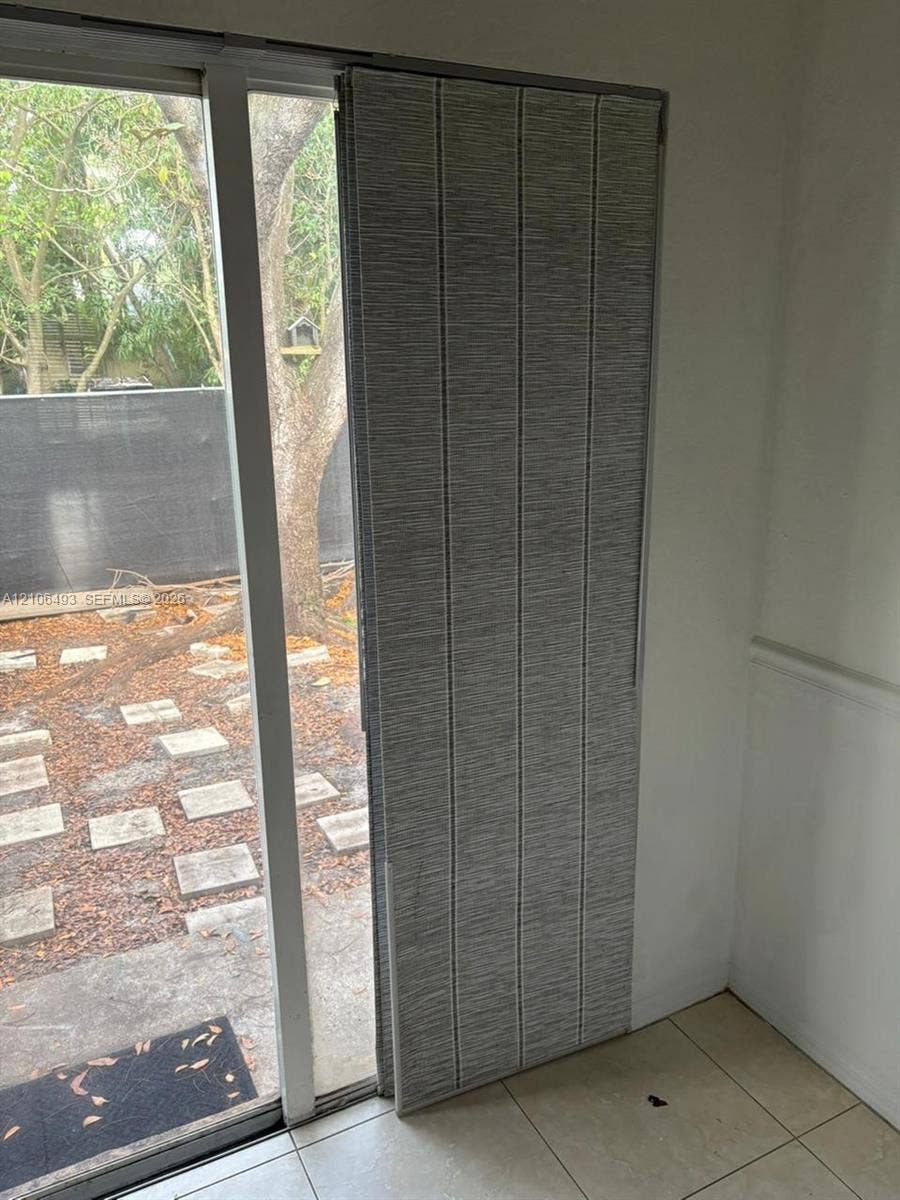 117 SE 3rd Avenue, #7, Hallandale Beach, FL 33009 Large-format glass sliding door with white frame and floor-to-ceiling textured panel blinds, featuring ceramic tile flooring and outdoor access to a patio with stone pavers