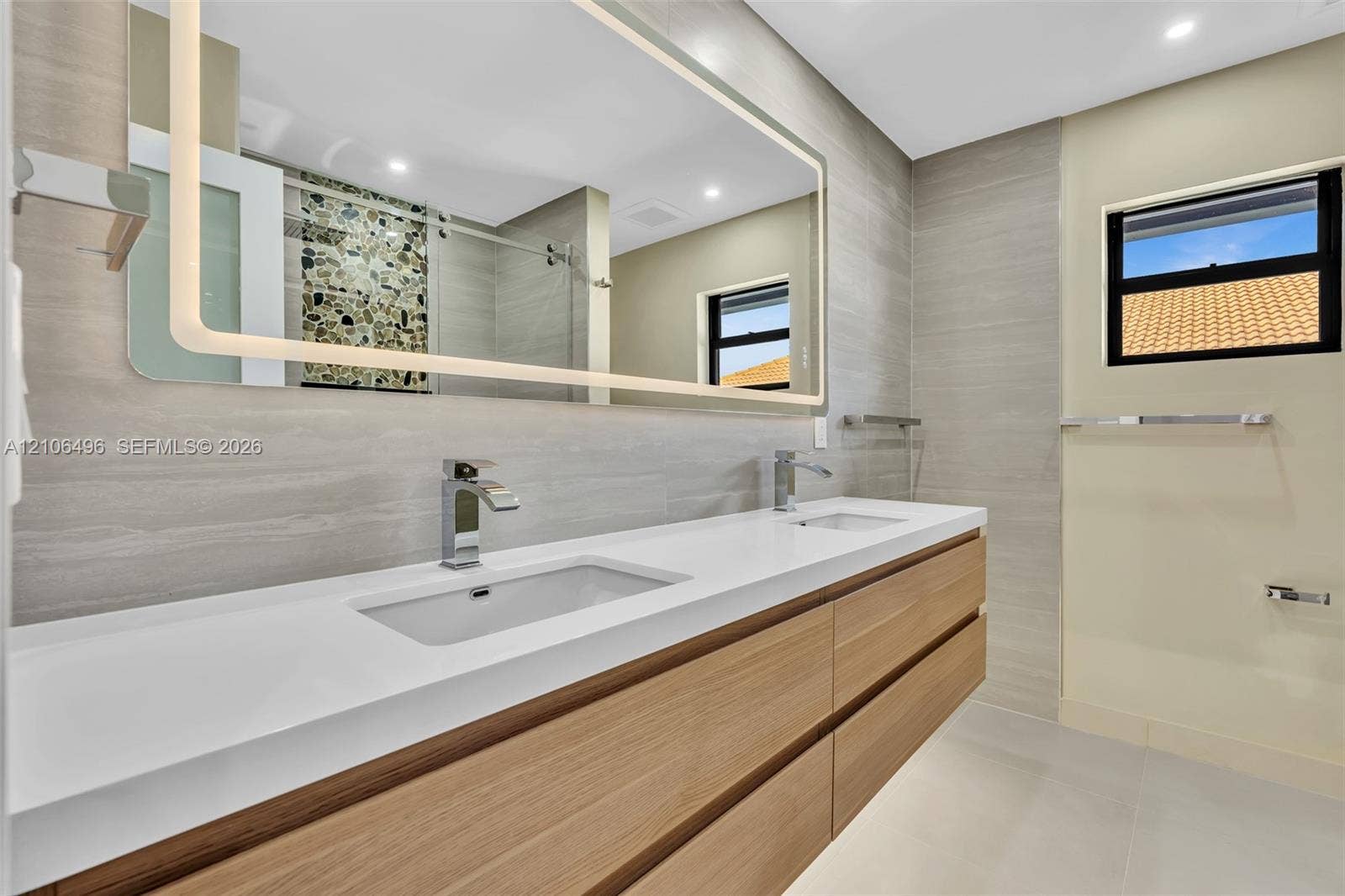 Double vanity featuring a white composite countertop, integrated rectangular sinks, wood-finish cabinetry, a large back-lit mirror, and neutral wall-tile surround
