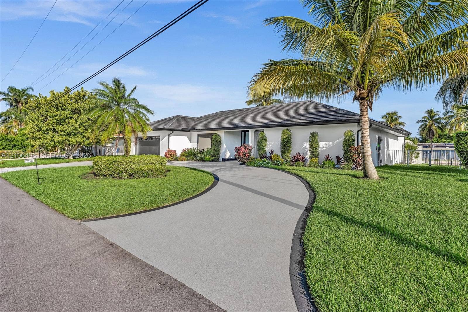 Single-story residence featuring a low-slope roof, white exterior cladding, a curved paved driveway, and established tropical landscaping