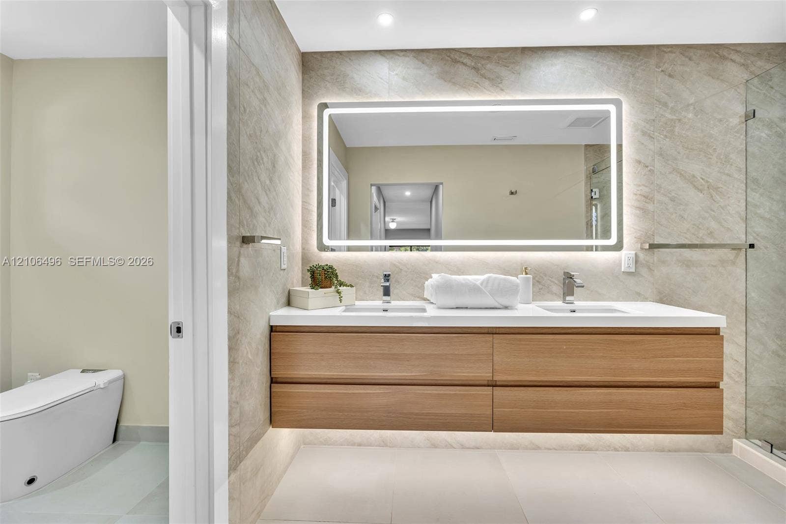 Floating wood-finish vanity with dual integrated sinks, quartz countertop, illuminated LED vanity mirror, full-height stone-finish wall tile, and glass-enclosed shower