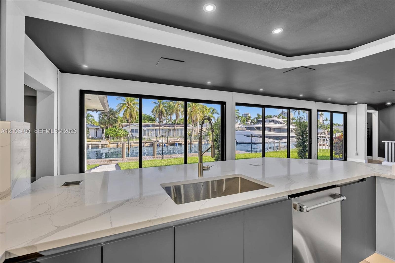 Large kitchen island with white countertop, undermount stainless steel sink, gooseneck faucet, gray flat-panel cabinetry, and extensive sliding glass doors