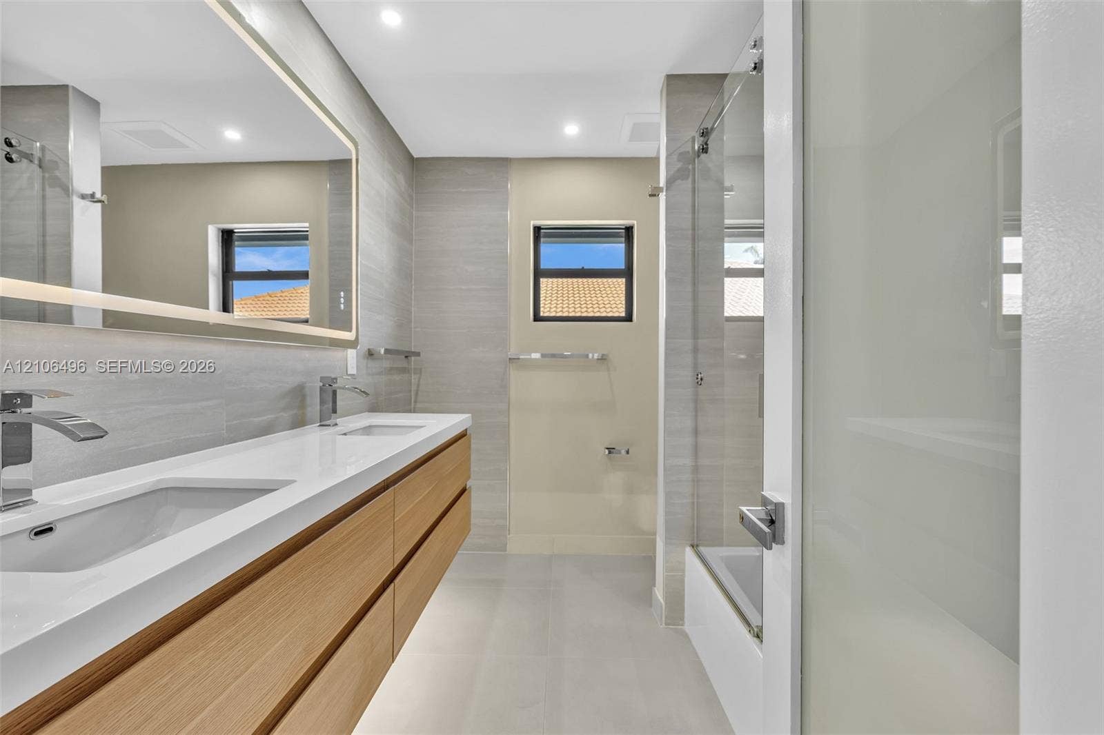 Floating double vanity with a stone countertop and dual undermount sinks, illuminated mirror, wood-finish cabinetry, and a glass-enclosed shower featuring textured wall tile