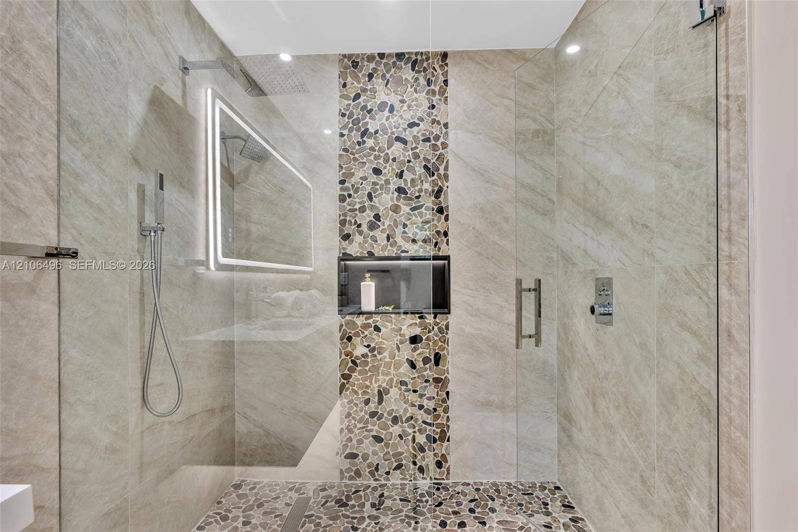 Walk-in shower featuring large-format neutral wall tile, a vertical pebble-stone accent column, pebble-tile flooring, a recessed storage niche, and an LED-backlit mirror