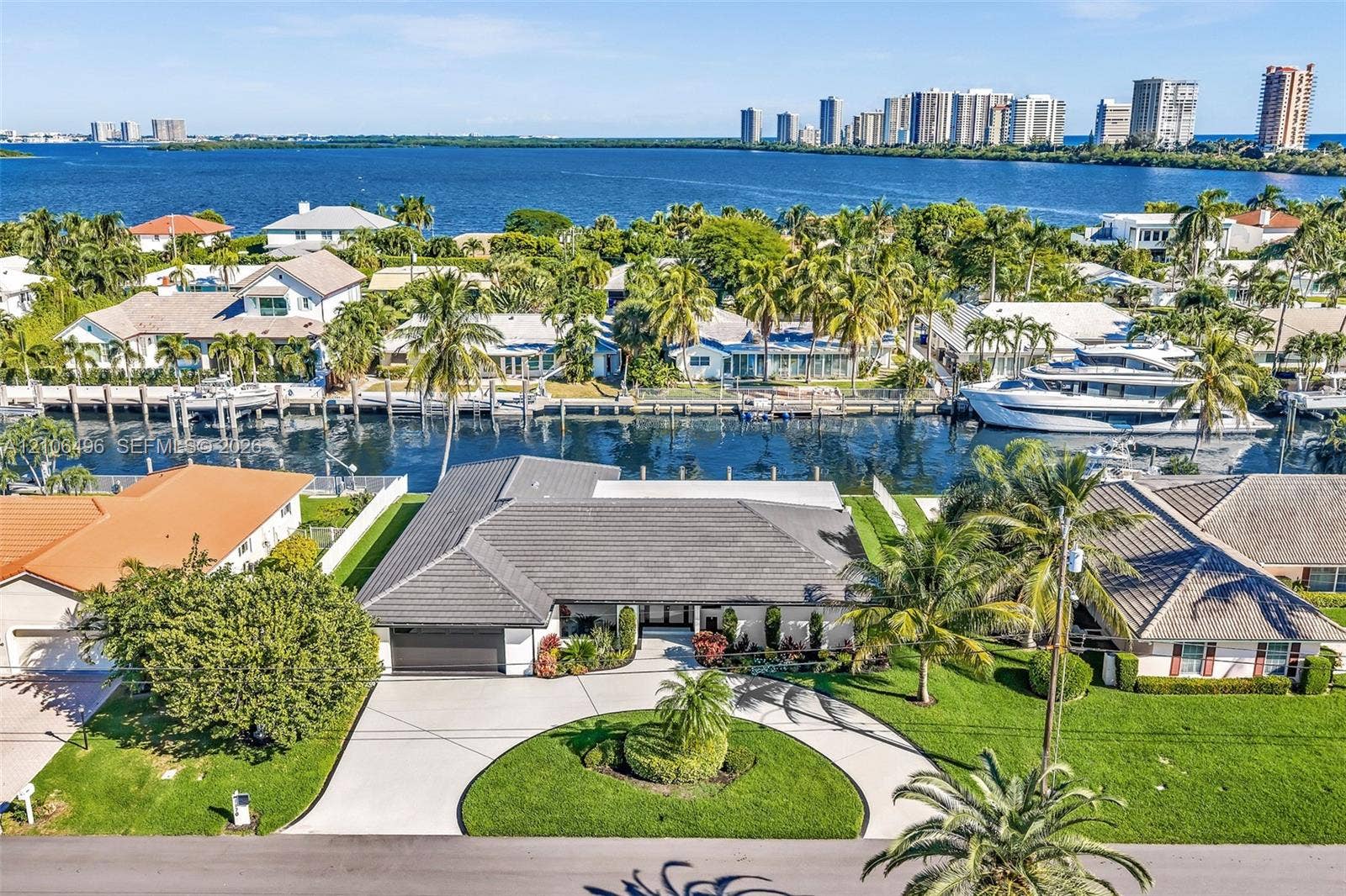Waterfront single-story residence featuring a tile roof, concrete circular driveway, manicured lawn, and mature palm trees