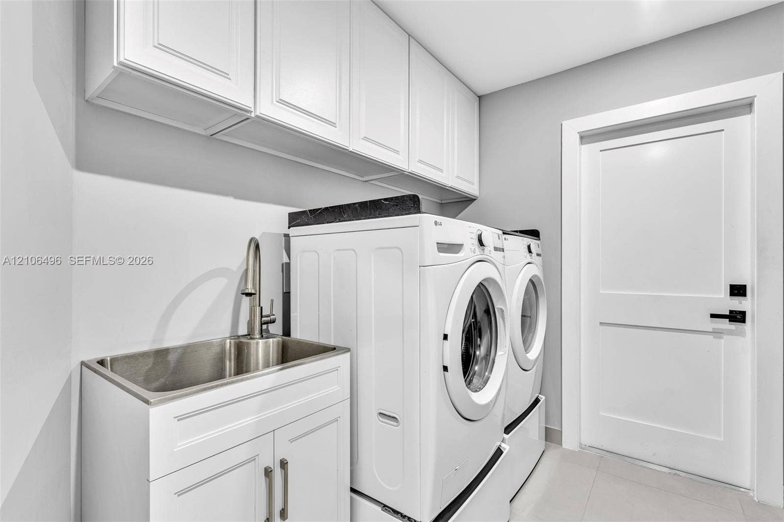 Laundry room featuring white shaker-style upper and lower cabinetry, an integrated stainless steel utility sink with a high-arch pull-down faucet, dark stone-finish countertop, and a solid-core door with matte black hardware