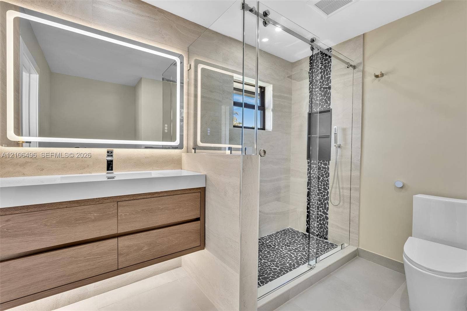 Floating wood-finish vanity with an integrated countertop, large LED-backlit mirror, and walk-in shower featuring glass enclosure, pebble stone flooring, and recessed shelving