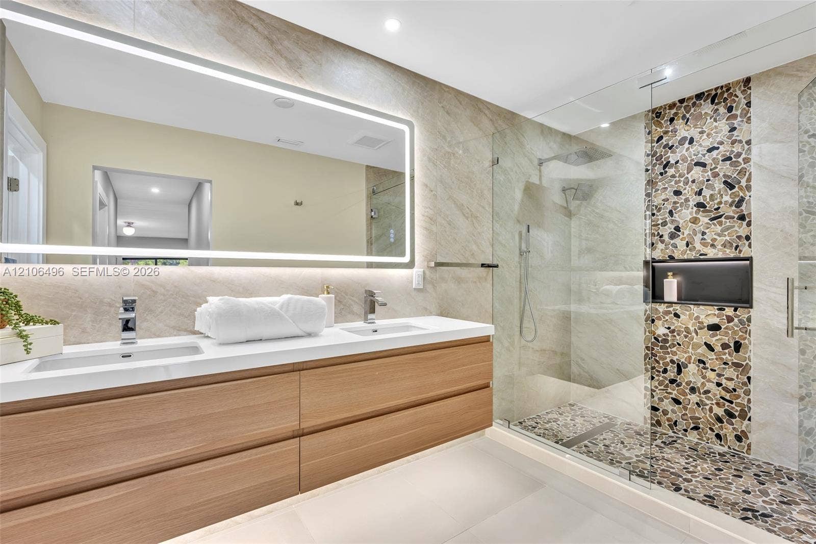 Floating wood-finish double vanity with rectangular integrated sinks, expansive LED-backlit mirror, and glass-enclosed shower featuring a river-stone floor and integrated wall niche