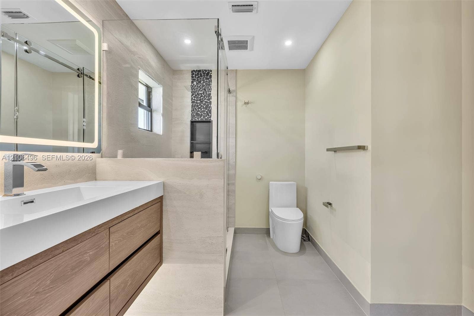 Floating wood-finish vanity with an integrated rectangular sink, backlit vanity mirror, glass-enclosed shower with decorative stone-look inlay, and large-format tile flooring