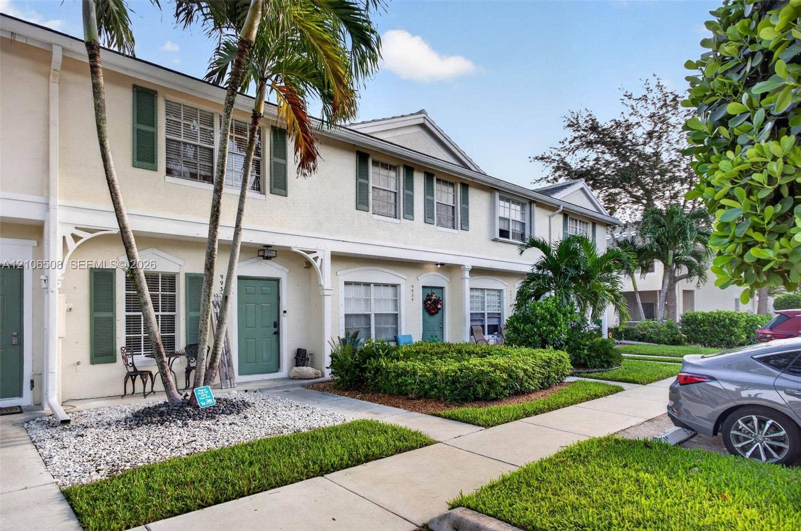 Stucco townhouse exterior featuring decorative shutters, covered entryways with arched brackets, and mature tropical landscaping