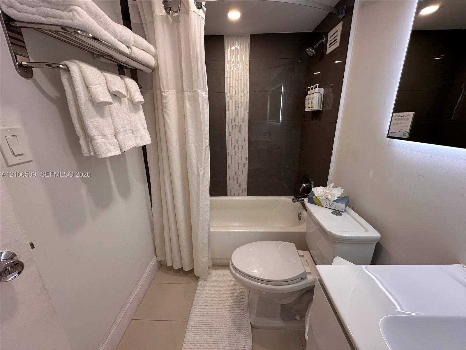Bathroom featuring a recessed tub with a vertical decorative tile inlay, wall-mounted towel rack, LED-backlit mirror, and neutral-toned floor tile