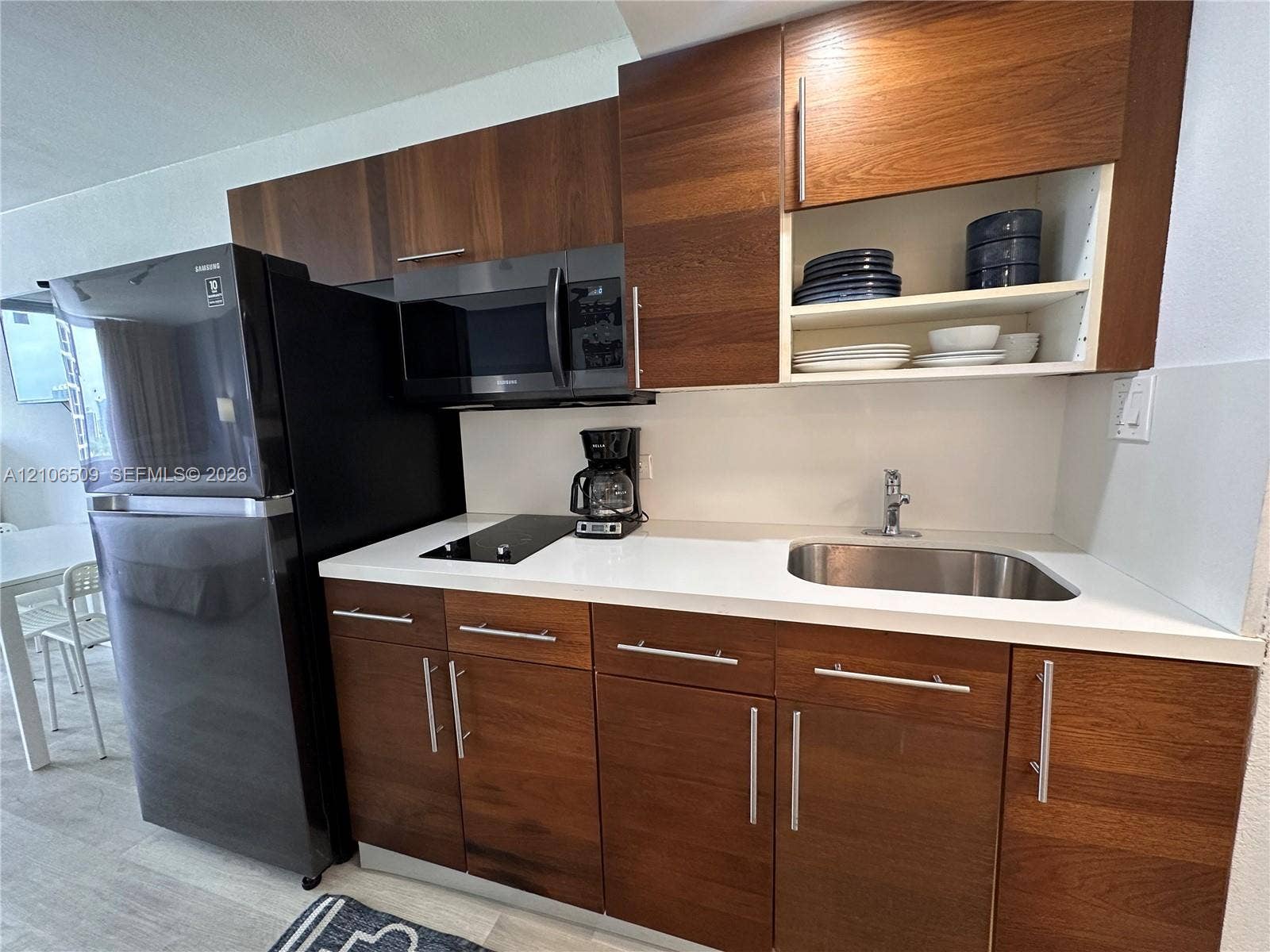 Kitchen featuring wood-finish flat-panel cabinetry, white solid-surface countertops, integrated stainless steel sink, black microwave, and black refrigerator