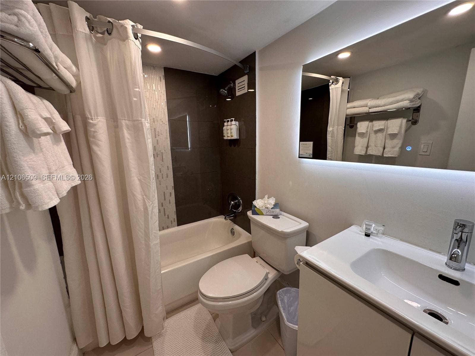 Bathroom featuring an integrated vanity with a rectangular sink, wall-mounted illuminated mirror, and a shower-tub combination with tiled surround and metal hardware
