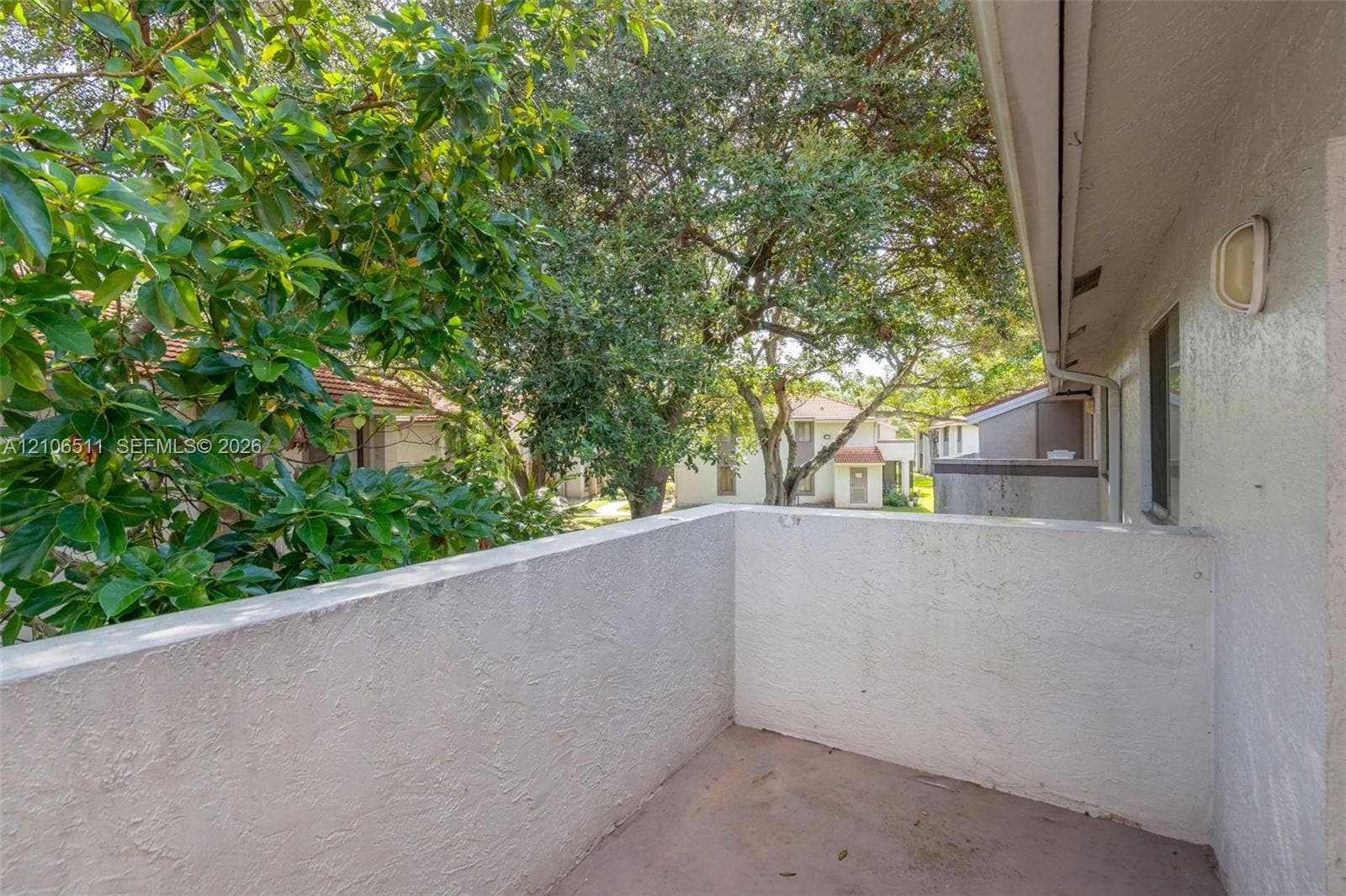 3229 Coral Lake Drive, #3229, Coral Springs, FL 33065 Covered balcony with textured stucco walls, solid wall railing, and an attached exterior light fixture, surrounded by mature trees