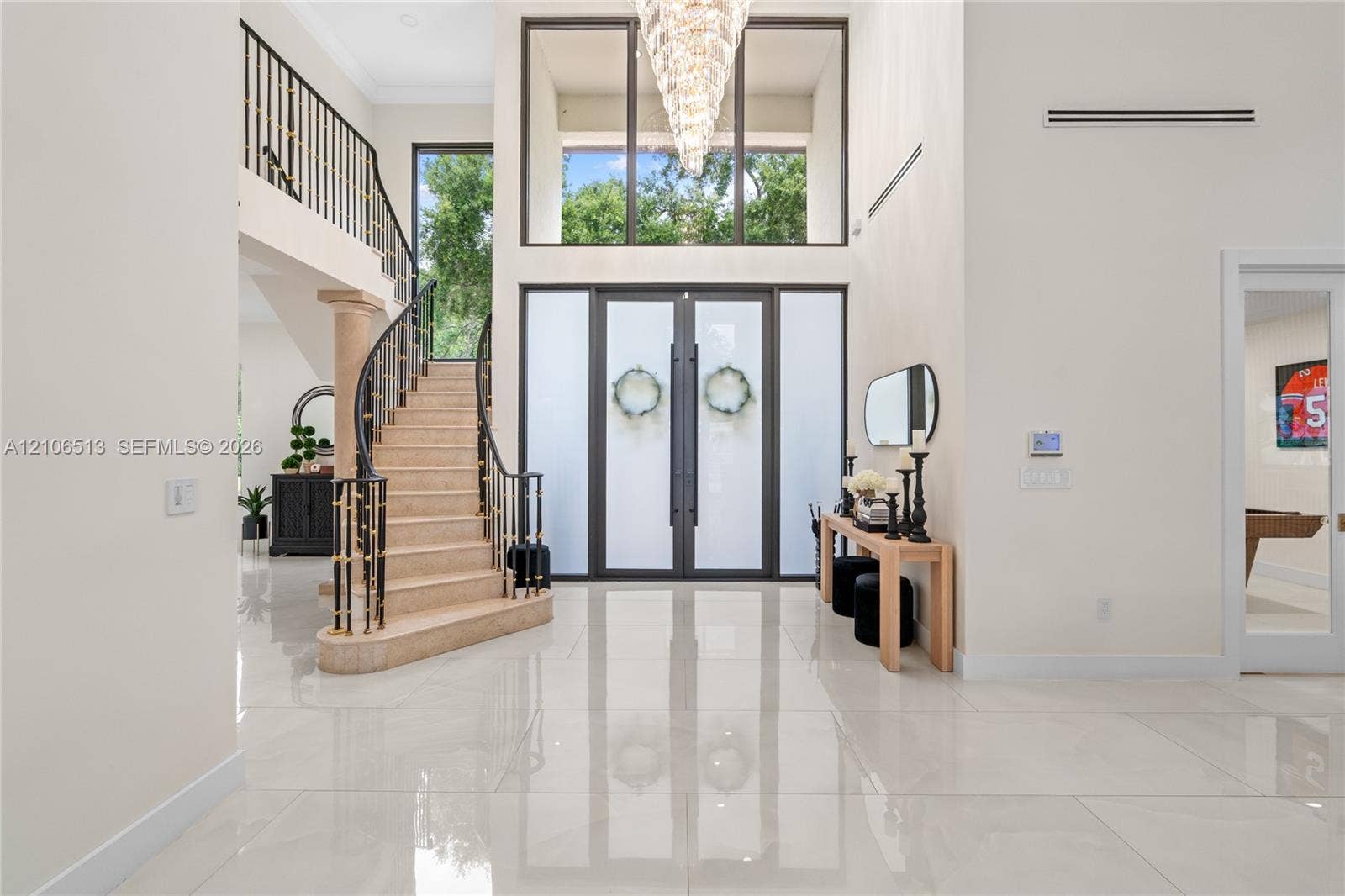 5720 SW 128th Street, Pinecrest, FL 33156 Grand foyer featuring polished large-format tile flooring, double entry doors with frosted glass panels, an expansive multi-story window, a tiered crystal chandelier, and a curved staircase with metal railings