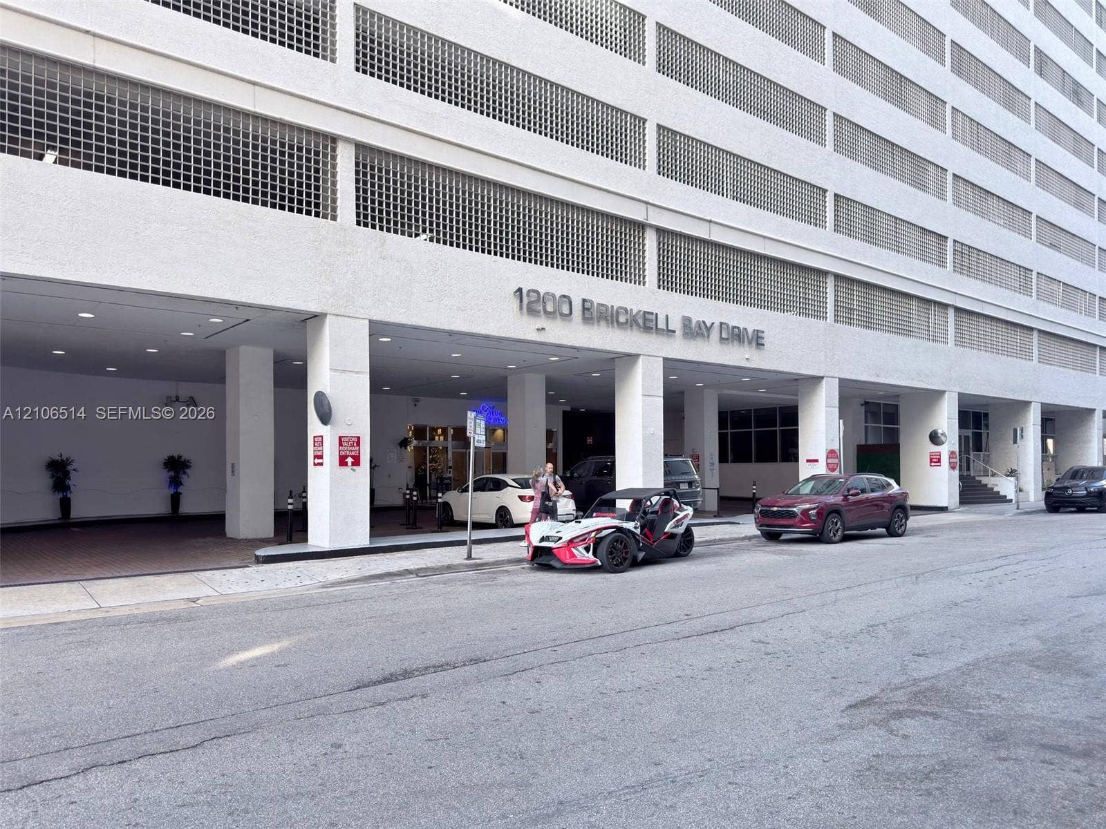Multi-story high-rise exterior featuring a recessed concrete porte-cochre, column-supported overhang, recessed ceiling lighting, and structured parking levels