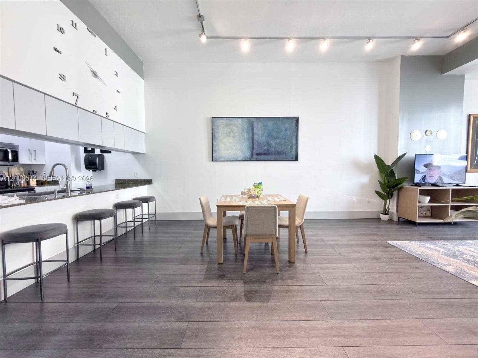 Open-concept interior featuring wood-finish flooring, track lighting, cabinetry with matte-finish upper doors, and a stone-surface breakfast bar