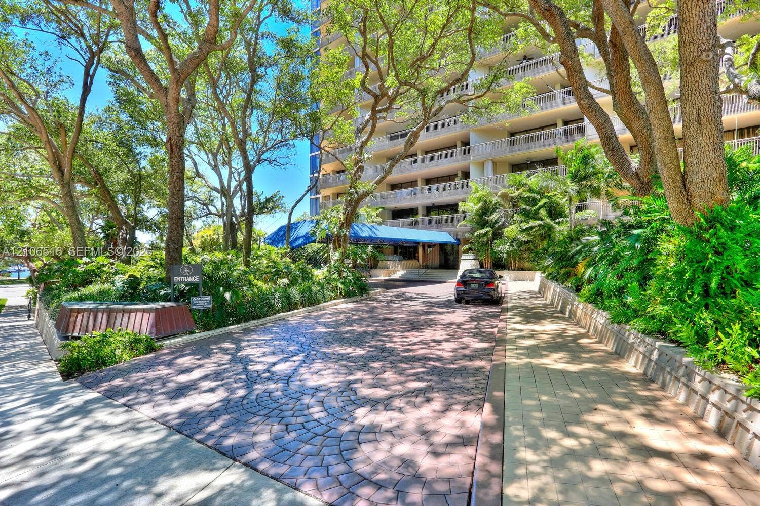Circular paved driveway featuring stamped pattern detailing, covered porte-cochre, multi-story building exterior, mature tree canopy, and landscaped greenery