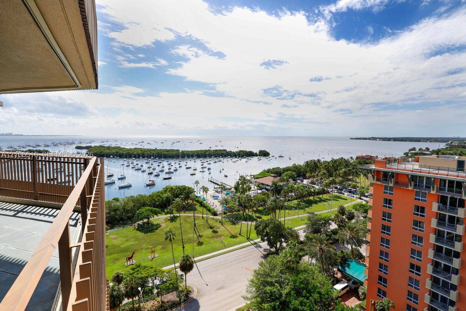 Elevated outdoor balcony with metal railing, overlooking a coastal expanse, harbor, and landscaped green space