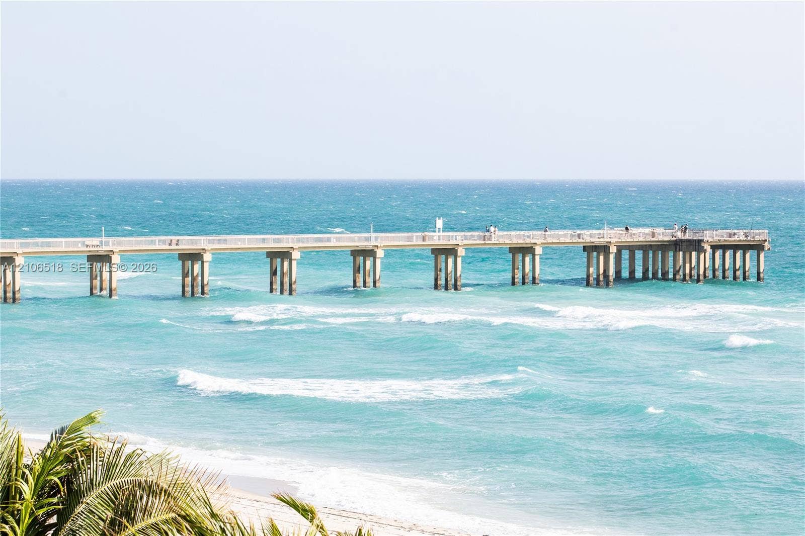 16500 Collins Avenue, #554, Sunny Isles Beach, FL 33160 Expansive pier with white structural railing extending over turquoise ocean waters, complemented by coastal sandy shoreline and tropical palm foliage