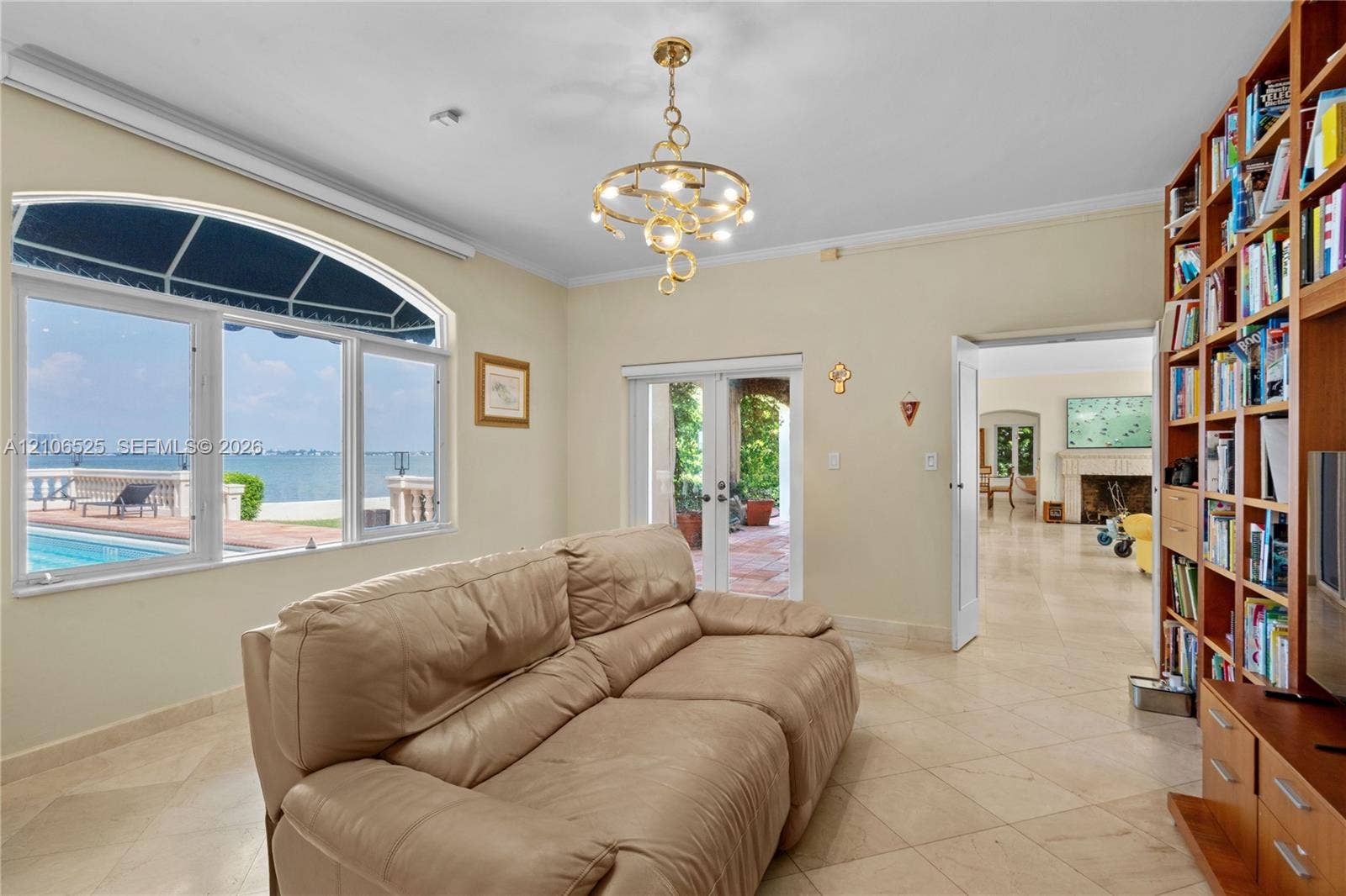 4760 N Bay Road, Miami Beach, FL 33140 Large arched window with water and pool expanse, built-in floor-to-ceiling wood shelving, gold-tone contemporary chandelier, and light-colored stone flooring