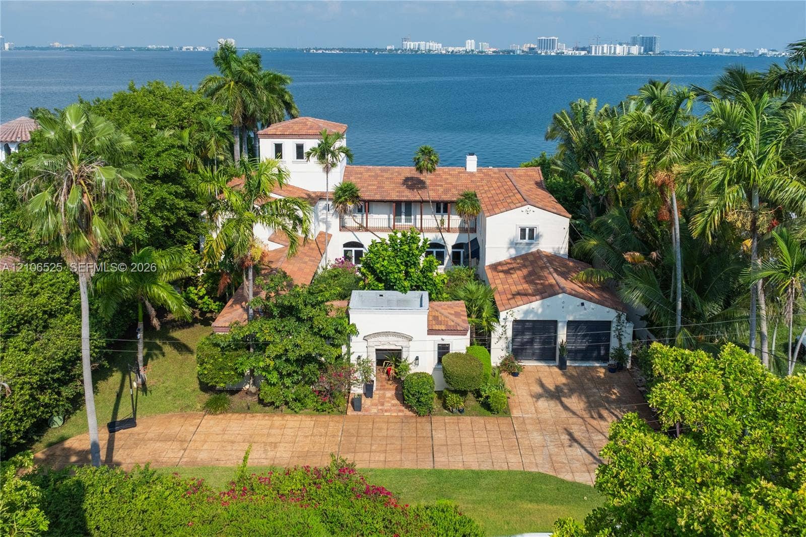 4760 N Bay Road, Miami Beach, FL 33140 Mediterranean-style estate featuring a terracotta tile roof, multi-story tower architecture, a paved motor court, and an attached two-car garage, situated on a waterfront lot with mature palm landscaping