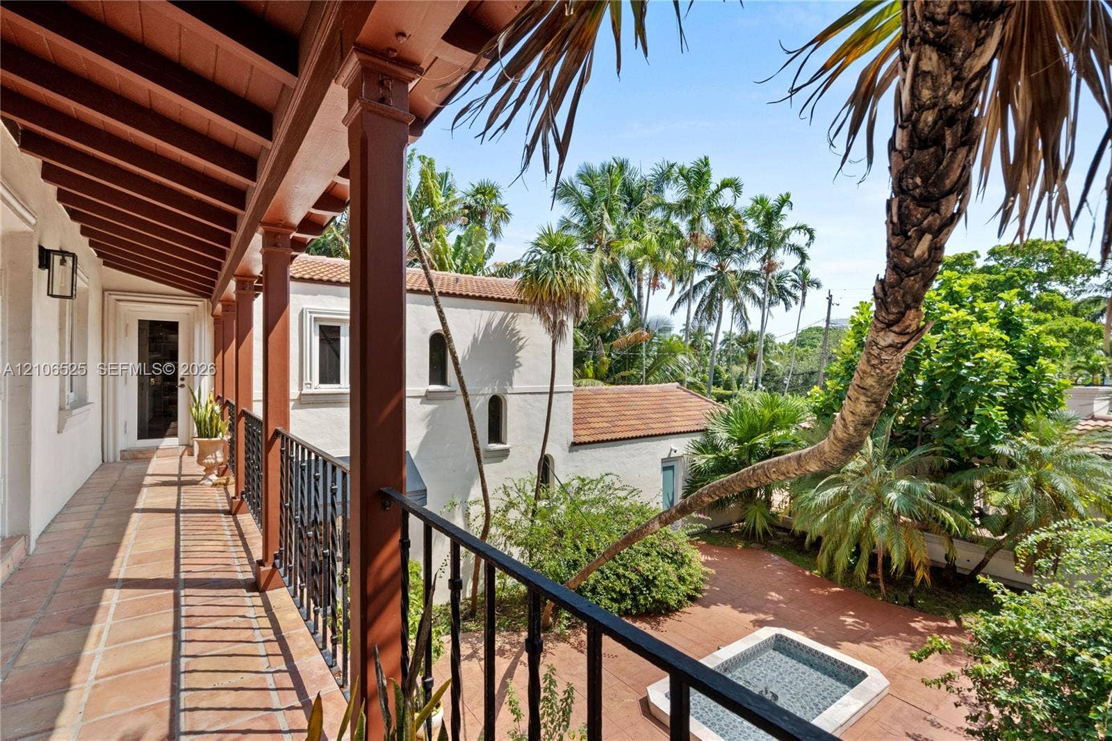 4760 N Bay Road, Miami Beach, FL 33140 Covered balcony featuring exposed wood beam ceilings, wrought-iron railings, and tile flooring, overlooking a paved courtyard with a built-in stone fountain