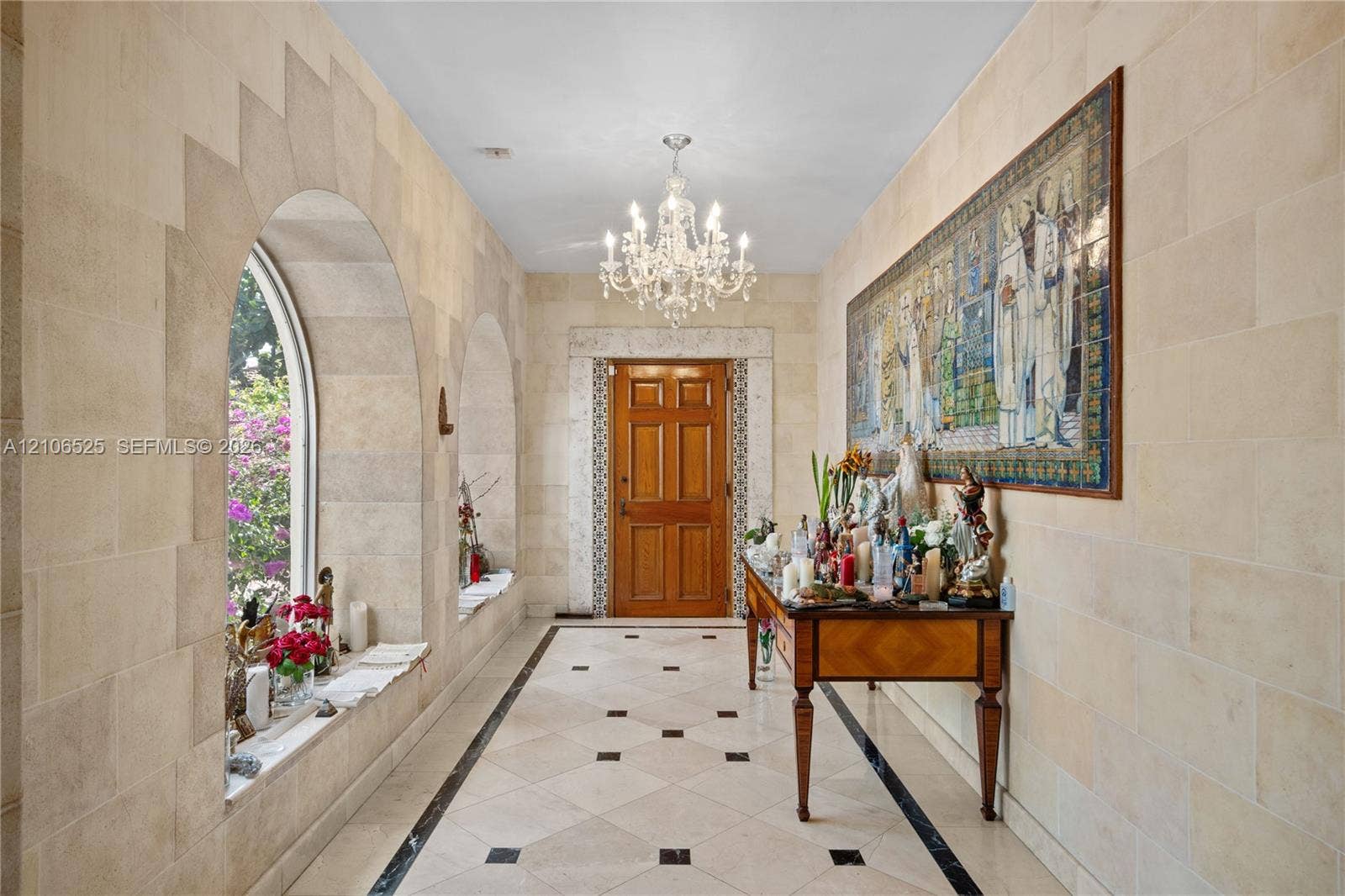 4760 N Bay Road, Miami Beach, FL 33140 Entryway featuring natural stone block walls, arched window alcoves, tile flooring with decorative stone insets, a crystal chandelier, and a solid wood door