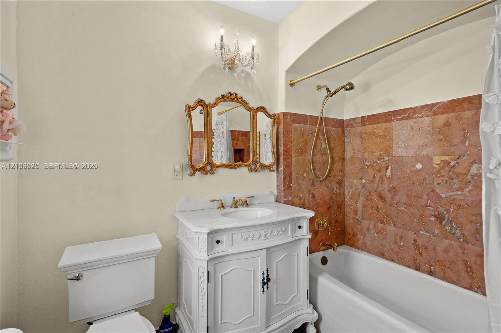 4760 N Bay Road, Miami Beach, FL 33140 Bathroom featuring a white vanity with carved detailing, stone tile shower surround, brass-finish fixtures, gold-toned ornamental mirror, and a crystal-accented wall-mounted light fixture