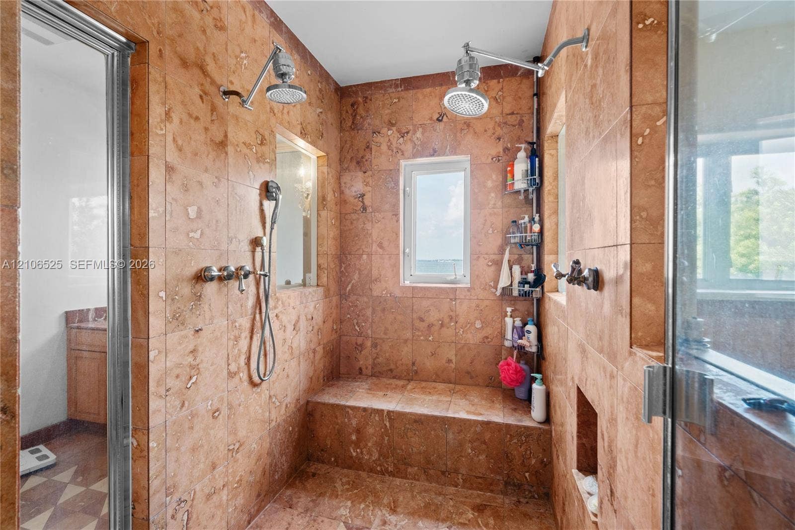 4760 N Bay Road, Miami Beach, FL 33140 Walk-in shower enclosure featuring floor-to-ceiling stone tile, a built-in tiled bench, dual overhead rainfall showerheads, a handheld shower fixture, and recessed wall shelving