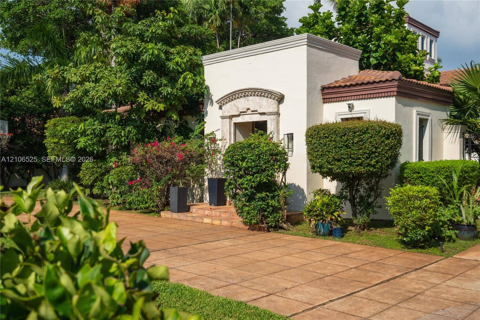 4760 N Bay Road, Miami Beach, FL 33140 Stucco exterior featuring arched stone entryway detail, tile roof, and expansive terracotta-style paved patio area surrounded by mature landscaping