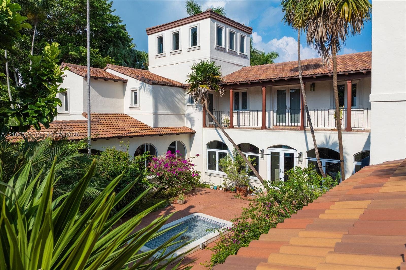 4760 N Bay Road, Miami Beach, FL 33140 Mediterranean-style exterior featuring a stucco facade, clay tile roof, central courtyard, second-story balcony with metal railing, and a built-in stone-accented fountain